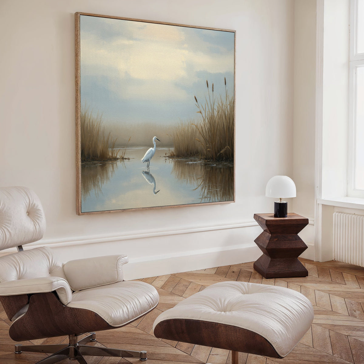 A modern living room with a white leather lounge chair and ottoman, a geometric side table and lamp, and Heron's Morning Canvas Art - Square depicting a serene egret scene displayed above the sofa.