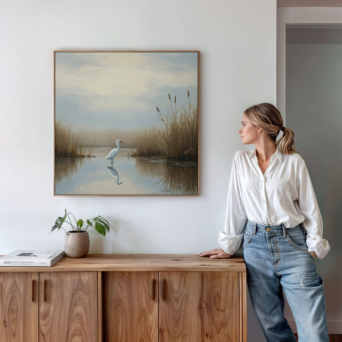 A woman in a white blouse and jeans stands beside a wooden cabinet, admiring the Heron's Morning Canvas Art - Square, which features a white egret in a serene wetland scene. A plant and books decorate the cabinet.