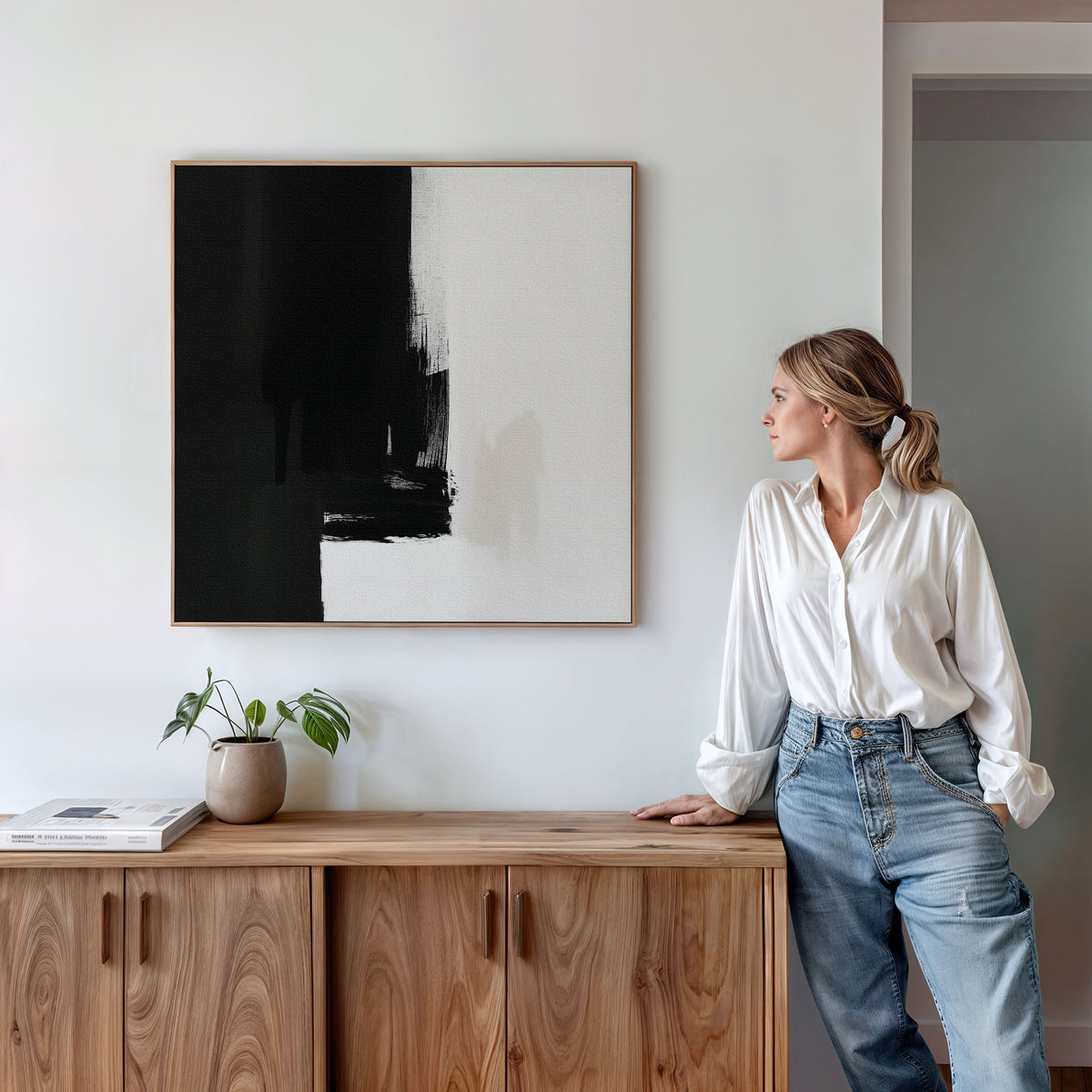 A woman in a white blouse and jeans leans on a wooden cabinet, gazing at the Ink Divide Canvas Art - Square above her. A potted plant and books on the cabinet enhance the serene, modern vibe of the space.