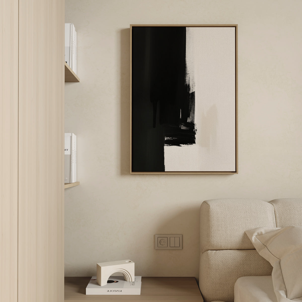 Minimalist living space with a beige sofa, light wood shelves holding books, and the Ink Divide Canvas Art - Vertical—a framed abstract black-and-white piece—accenting the cream wall. A small modern decorative object sits on the lower shelf.