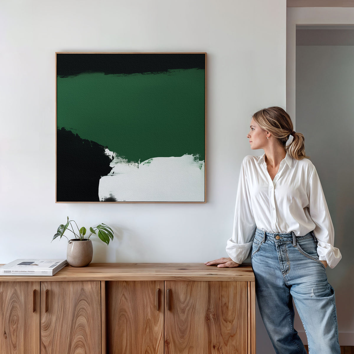 A woman in a white shirt and jeans stands by a wooden sideboard, admiring the Jade Depths Canvas Art - Square, an emerald green abstract piece that adds modern style to the living room. A small plant and book sit on the sideboard beneath the artwork.