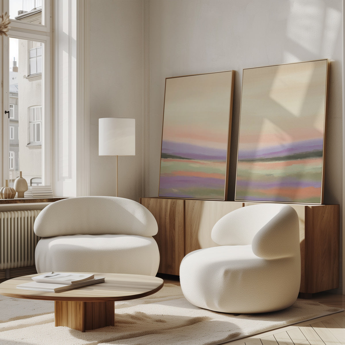 A modern living room with two rounded white armchairs, a low wooden coffee table, a tall floor lamp, and serene wall art—Lavender Valley - Set Of 2 pastel abstract paintings—leaning against a wooden sideboard beneath large windows.