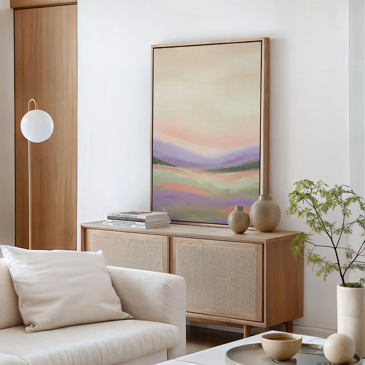 A modern living room with a light wood sideboard, the Lavender Valley Canvas Art - Vertical above, neutral vases, a stack of books, a white globe lamp, a white sofa, and a small potted plant on a white table.