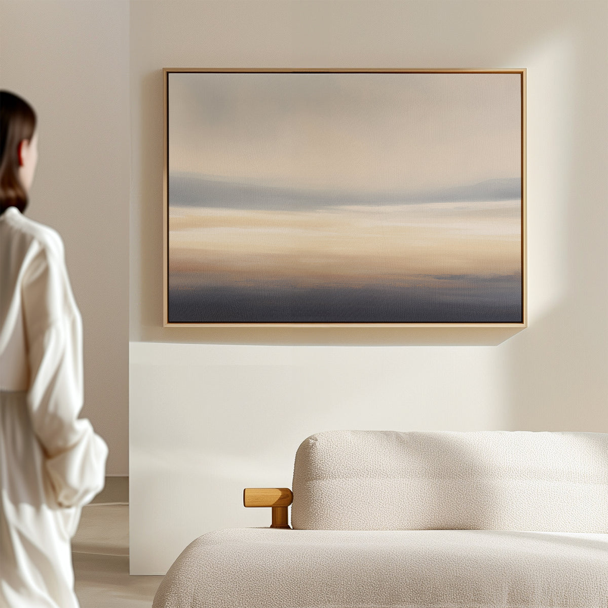 In a minimalist room, a person admires the Layered Dusk Canvas Art - Horizontal, featuring calming blue, beige, and gray tones inspired by Nordic Park, displayed above a light sofa on a cream-colored wall.