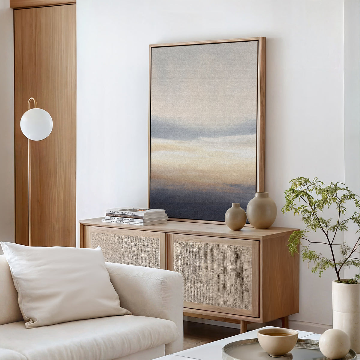 A minimal, modern living room with a light wood sideboard, Layered Dusk Canvas Art - Vertical, neutral vases, books, a potted plant, and a white sofa with a cushion. Soft natural light fills the space.
