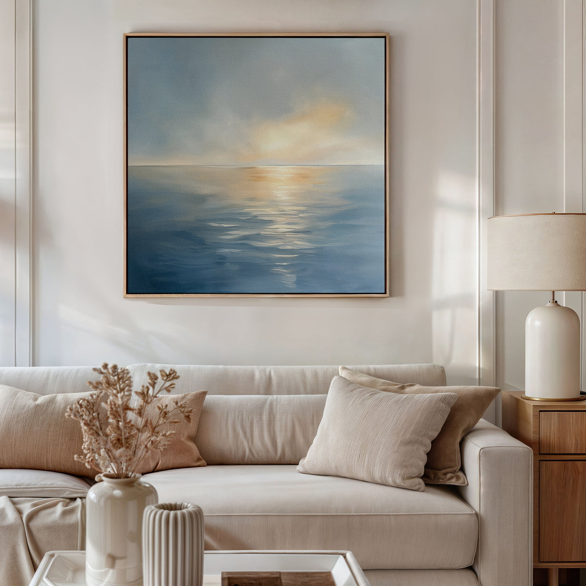 A modern living room features a beige sofa, neutral cushions, a table with dried florals, a white lamp, and the Luminous Calm Canvas Art - Square, adding a serene coastal palette to the space.