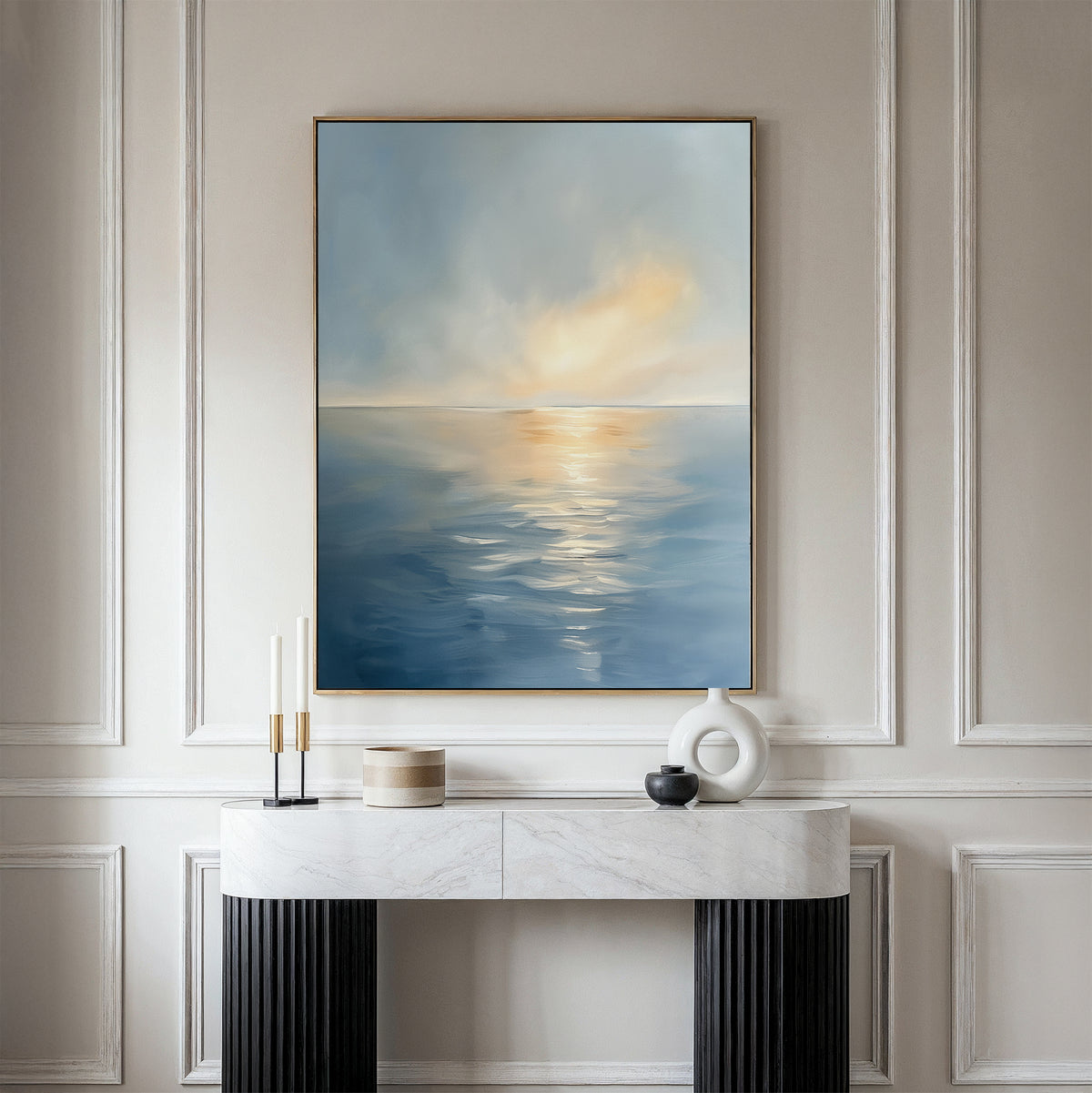 The Luminous Calm Canvas Art - Vertical, featuring a serene seascape in soft blue and orange tones, hangs on a white paneled wall above a marble console table decorated with modern accents and candles.
