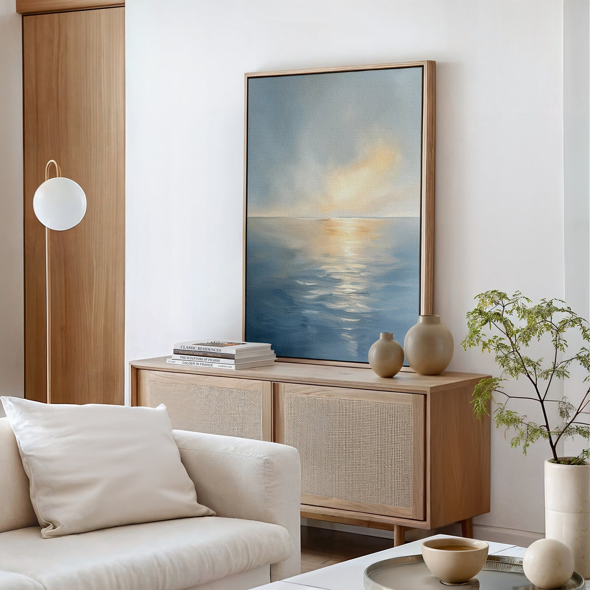 A serene living room with a wooden sideboard, the Luminous Calm Canvas Art - Vertical, neutral ceramics, a potted plant, globe floor lamp, and a white sofa with cushion create a minimalist space inspired by coastal tones.