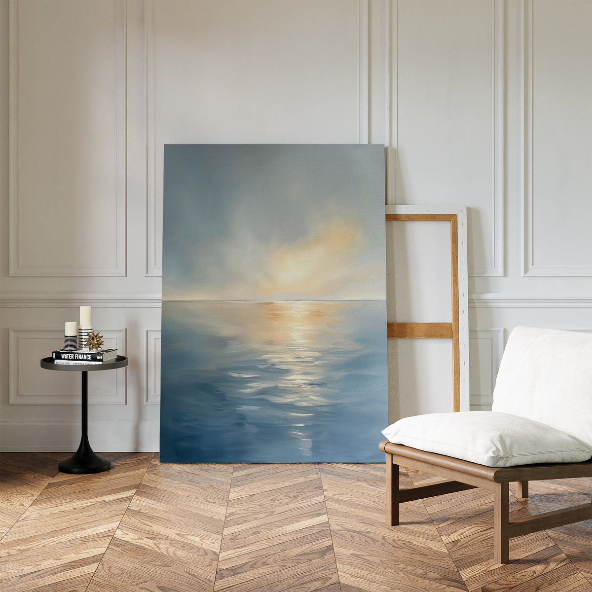 The Luminous Calm Canvas Art - Vertical, featuring a tranquil sea and soft sky in coastal shades, rests against a white paneled wall beside an empty frame, near a white cushioned chair and black side table with candles on herringbone wood flooring.