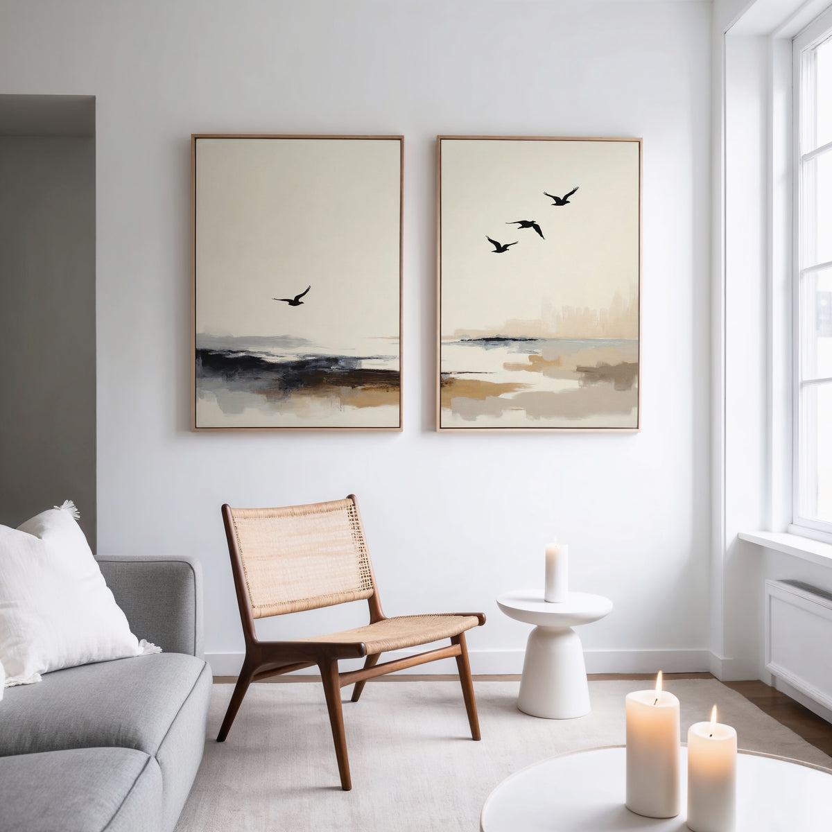 A modern living room with a gray sofa, a woven wooden chair, a small white side table with a lit candle, and the Maritime Flight - Set Of 2 coastal prints on the wall. Sunlight streams in through large windows.