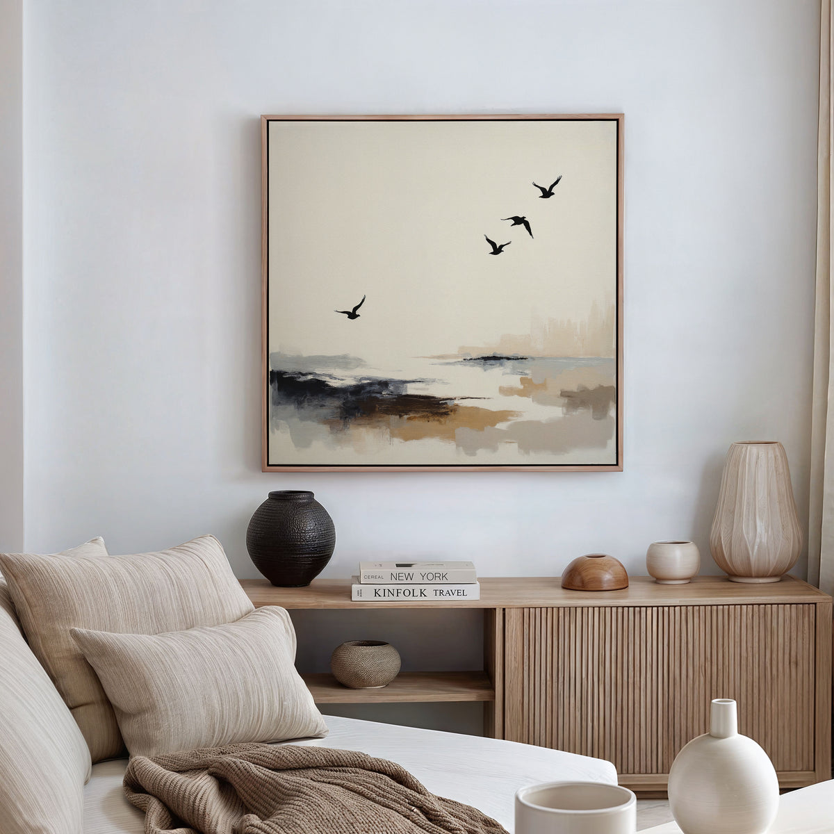 A minimalist living room with neutral tones features a beige sofa, wooden sideboard with ceramic vases and books, and the Maritime Flight Canvas Art - Square—a modern coastal painting of flying birds adding an airy touch to the space.
