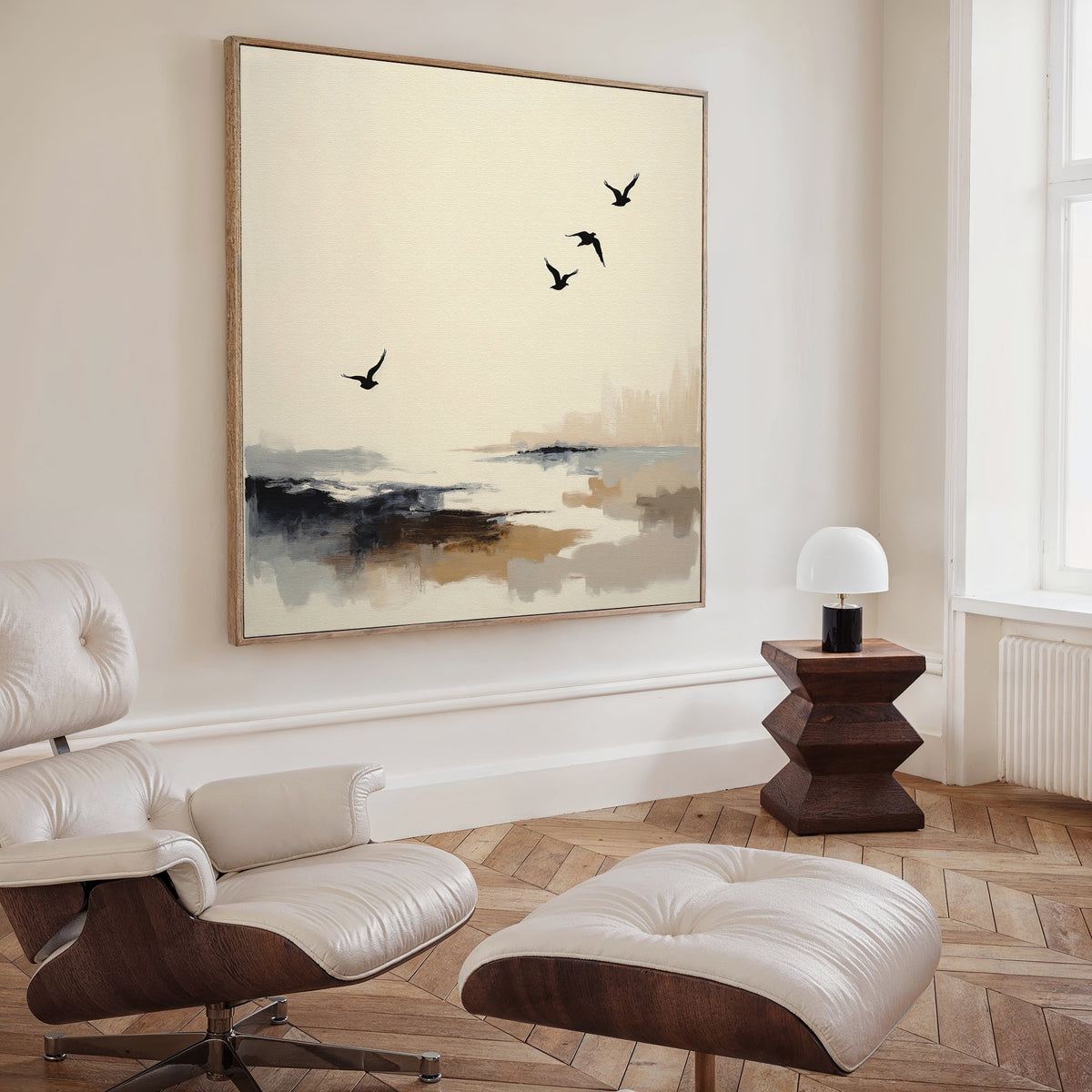 A modern, minimalist living room features a white lounge chair, ottoman, geometric wooden side table, small white lamp, and the Maritime Flight Canvas Art - Square, adding subtle nautical decor with birds soaring over an abstract coast.