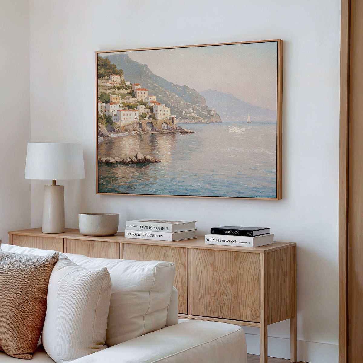 A cozy living room features a wooden sideboard with stacked books, a lamp, a bowl, and serene decor highlighted by the Mediterranean Dreams Canvas Art - Horizontal depicting a coastal village. A cream sofa is partially visible in front.