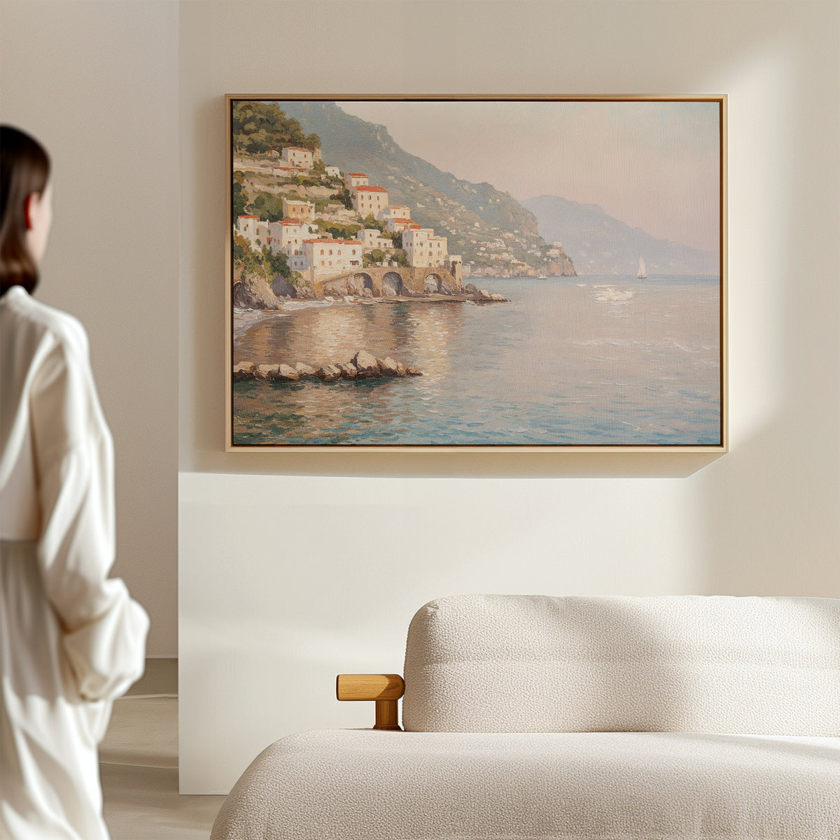 A person stands in a softly lit room, admiring the Mediterranean Dreams Canvas Art - Horizontal, which portrays a coastal village with colorful cliffside houses and calm blue water. A beige sofa in the foreground enhances the serene décor.