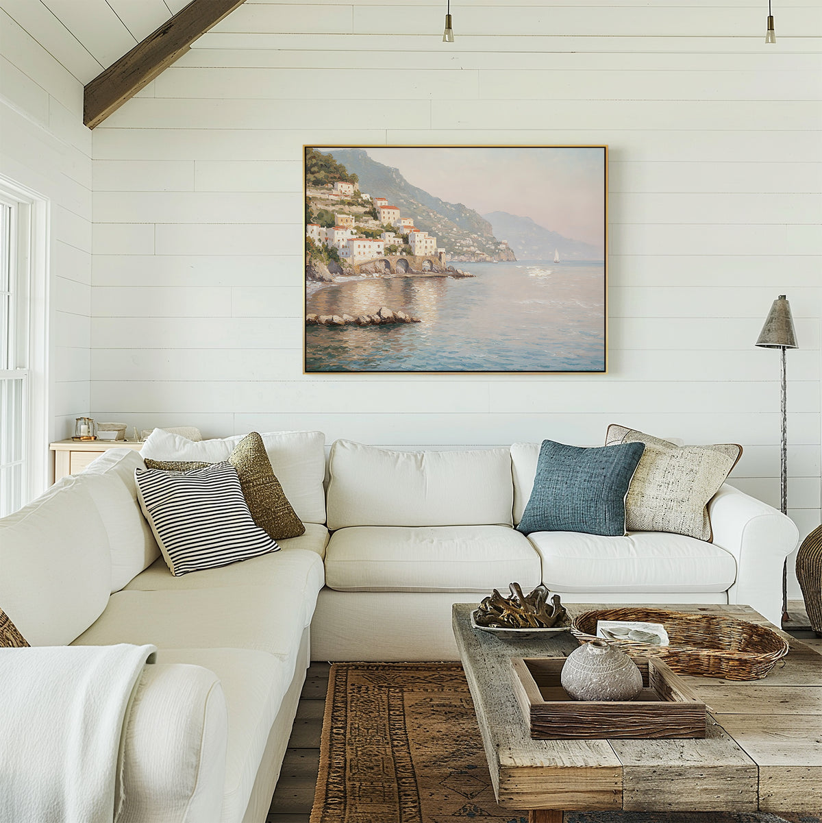 A cozy living room with a white sectional sofa, assorted pillows, a rustic wooden coffee table, and the Mediterranean Dreams Canvas Art - Horizontal depicting a coastal village on a white shiplap wall.