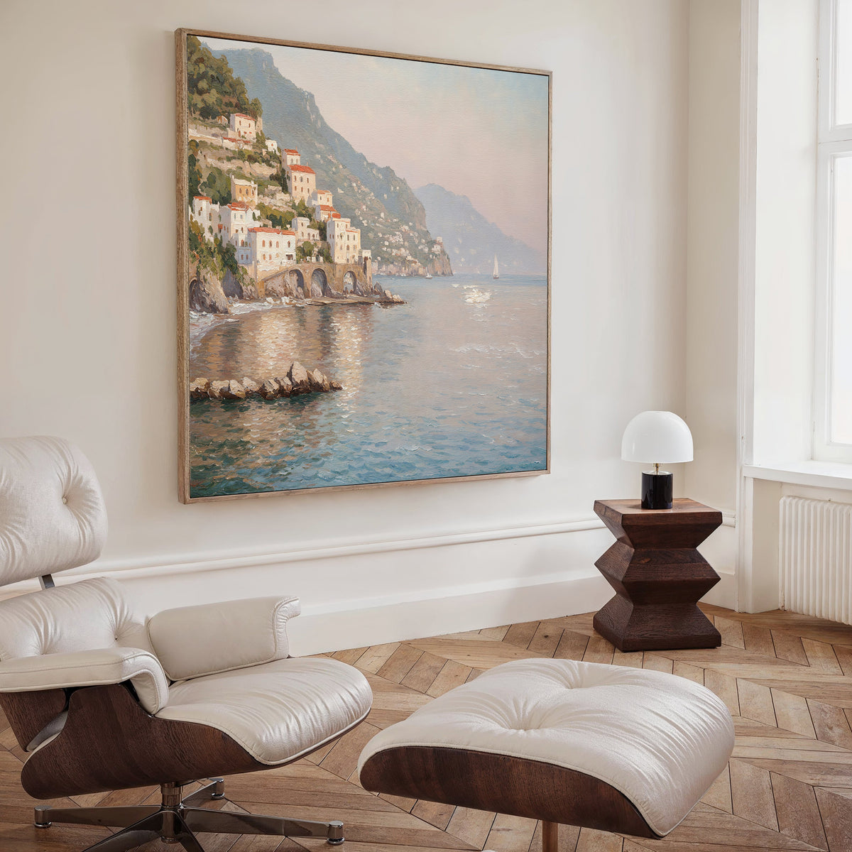 A cozy room features a white lounge chair and ottoman, a wooden side table with a modern lamp, and the Mediterranean Dreams Canvas Art - Square—a coastal village scene with mountains and calm water. Sunlight streams in, creating serene decor.