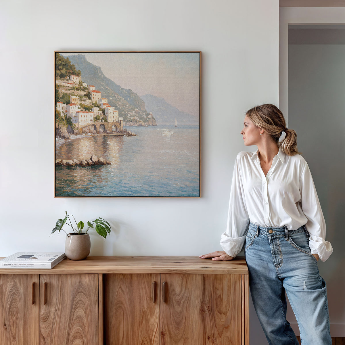 A woman in a white blouse and jeans leans on a wooden cabinet, admiring the Mediterranean Dreams Canvas Art - Square, which brings serene decor to the white wall. A potted plant and books complete the peaceful scene.