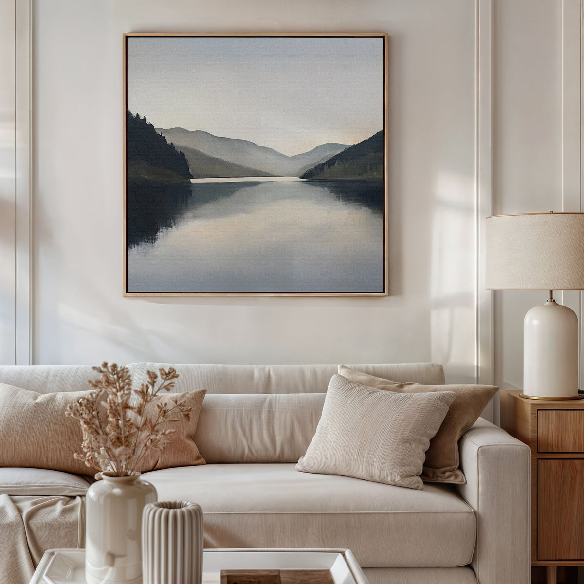 A cozy living room with a beige sofa, cream cushions, a side table lamp, and dried flowers in a vase. Above the sofa hangs the Mirror Valley Canvas Art - Square, showcasing a tranquil lake and mountain scene beneath a gentle sky.