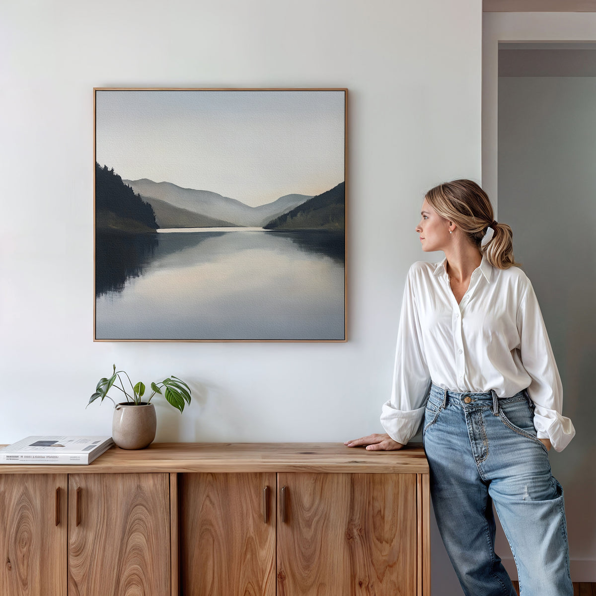 A woman in a white blouse and jeans leans against a wooden cabinet, admiring the Mirror Valley Canvas Art - Square hanging above a small plant and books on the white wall.