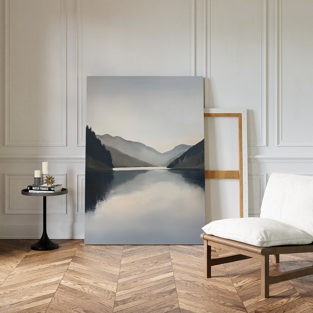 A minimalist room with wood herringbone floors, white walls, Nordic decor, a cushioned chair, a small round table with candles and books, and the Mirror Valley Canvas Art - Vertical of mountains and a lake resting against the wall.