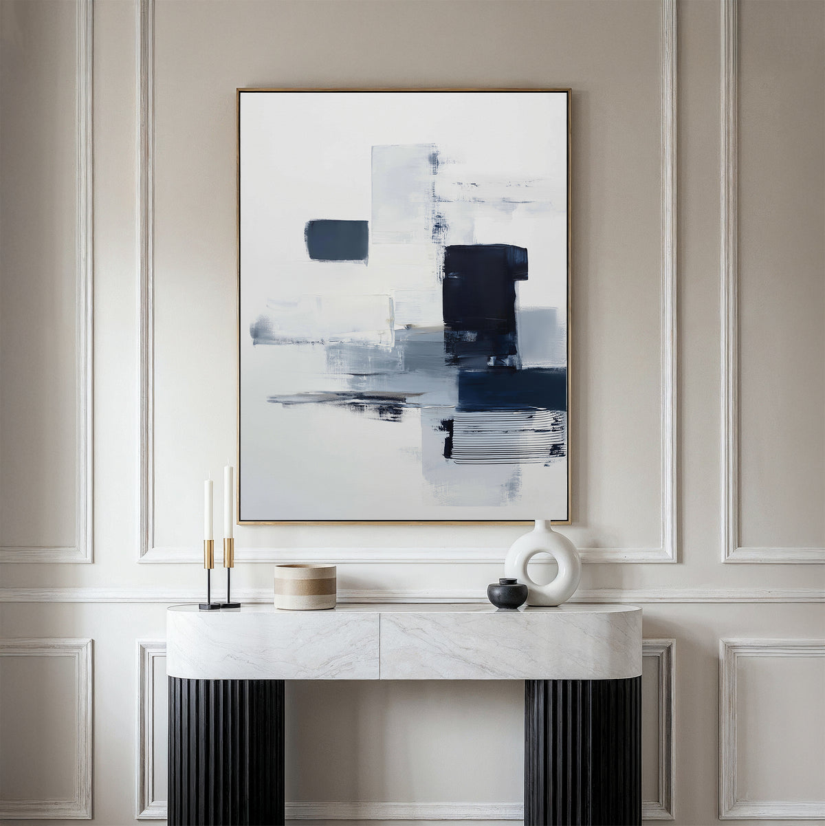 A modern, minimalist entryway includes a marble console table with black legs and decorative objects, highlighted by the Navy Brushwork Canvas Art - Vertical in blue, white, and black on a paneled cream wall.