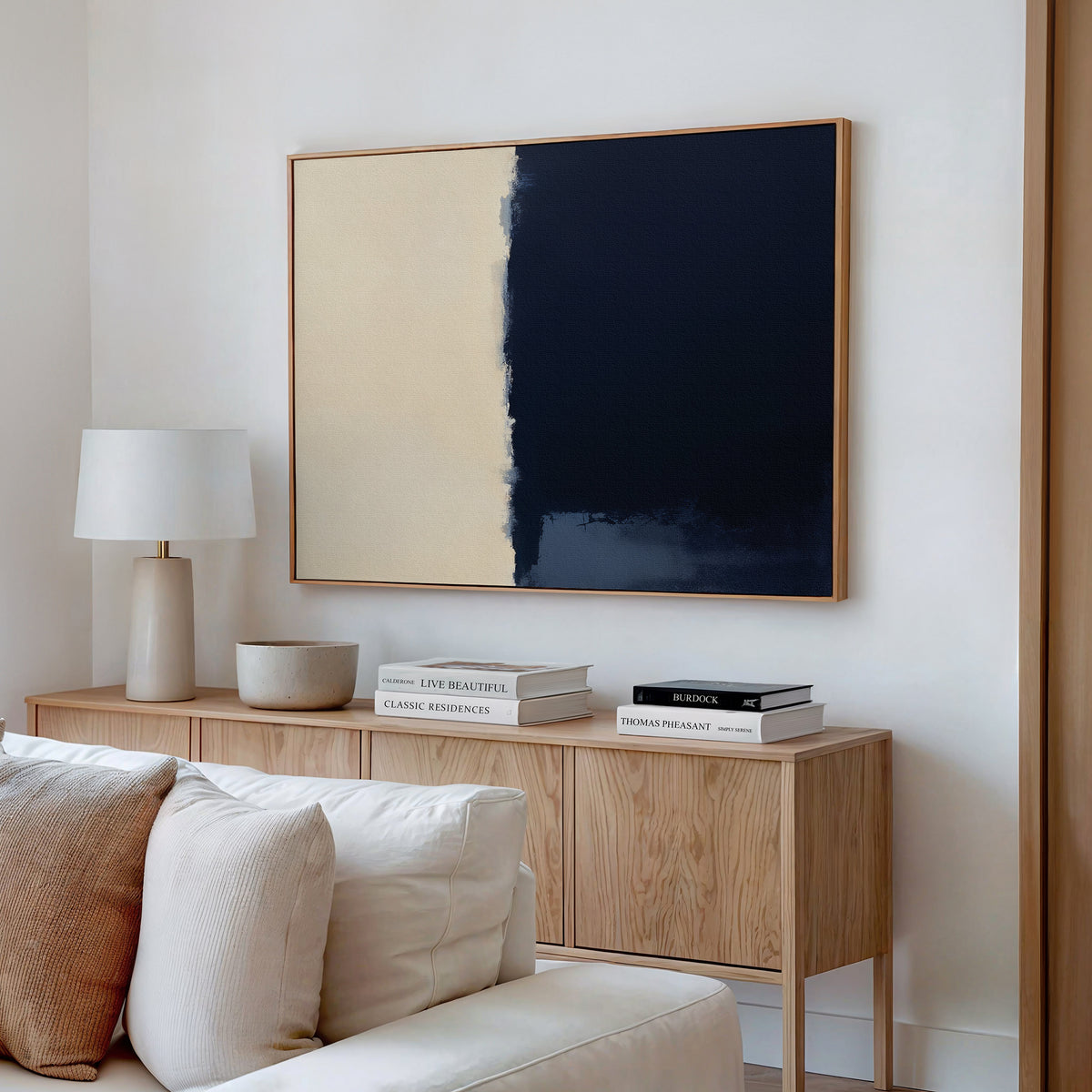 A minimalist living room features a light wood sideboard, stacked books, a white lamp, a bowl, and the Navy Division Canvas Art - Horizontal with beige and dark blue sections, adding modern elegance to the white wall.