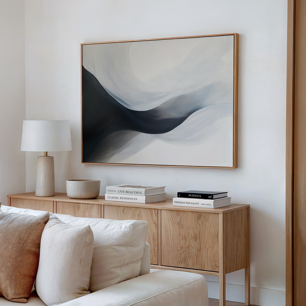 A modern living room with a light wood sideboard, beige sofa, table lamp, books, and the Rippling Vista Abstract Canvas Art in gray and white above the sideboard creates an atmosphere of modern elegance.