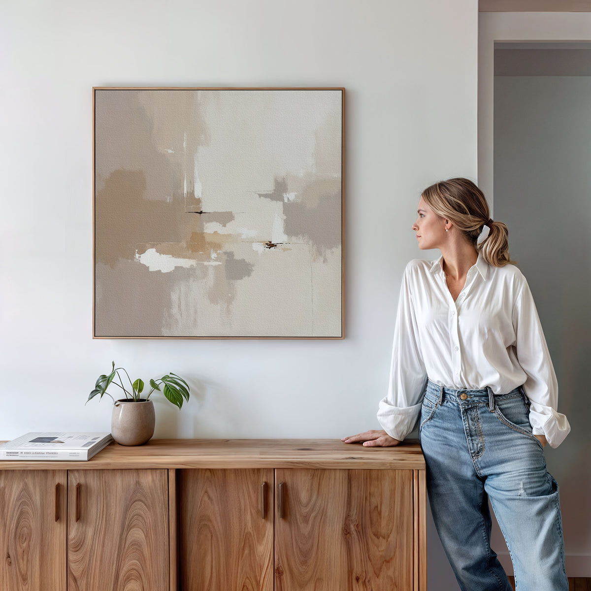 A woman in a white shirt and jeans leans against a wooden cabinet, admiring the Neutral Territory Canvas Art - Square on the wall, while a small plant and books add modern, minimalist style to the contemporary room.