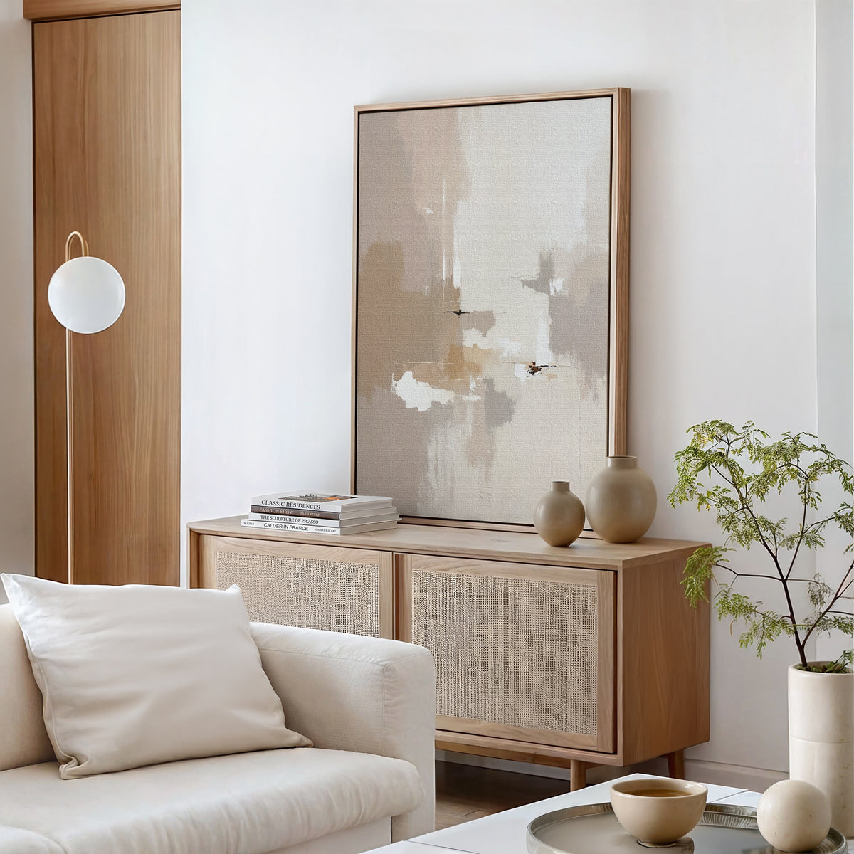 Minimalist living room featuring a light wood sideboard, Neutral Territory Canvas Art - Vertical, neutral vases, stacked books, a potted plant, white armchair with cushion, and round floor lamp beside a wooden door—ideal for modern spaces.