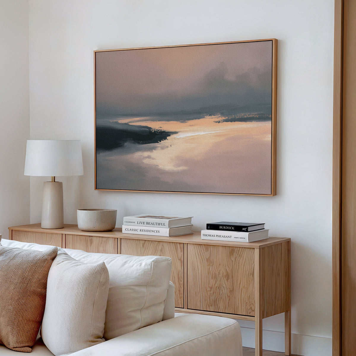 A modern living room with a light wood sideboard, books, lamp, and ceramic bowl. Above a cream sofa, the Nordic Fjord Canvas Art - Horizontal hangs on the white wall, featuring a warm, muted landscape print.