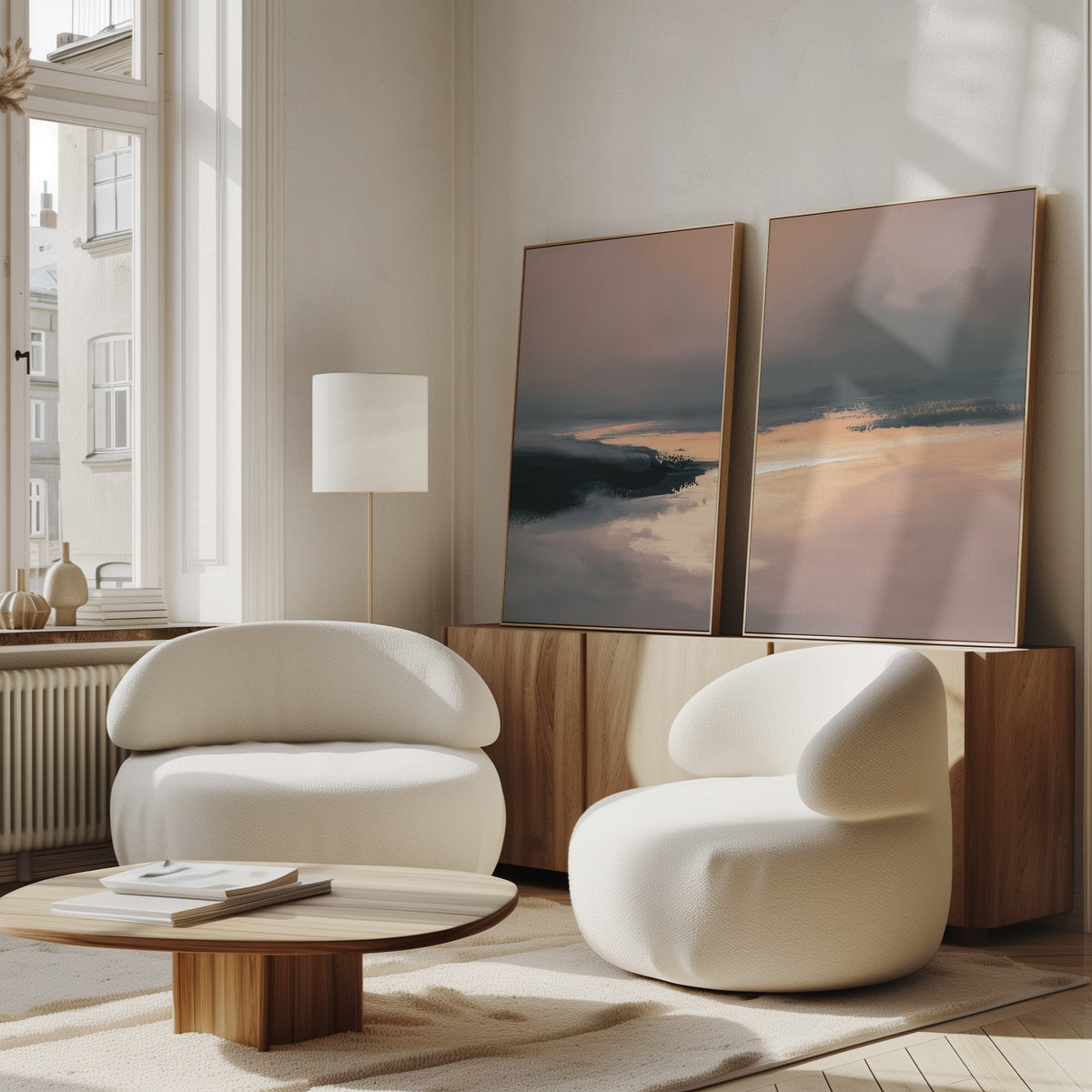 A modern living room features two curved white armchairs, a low wood coffee table, and a sideboard. The peaceful Nordic Fjord - Set Of 2 wall art leans against the white wall, illuminated by sunlight from tall windows.