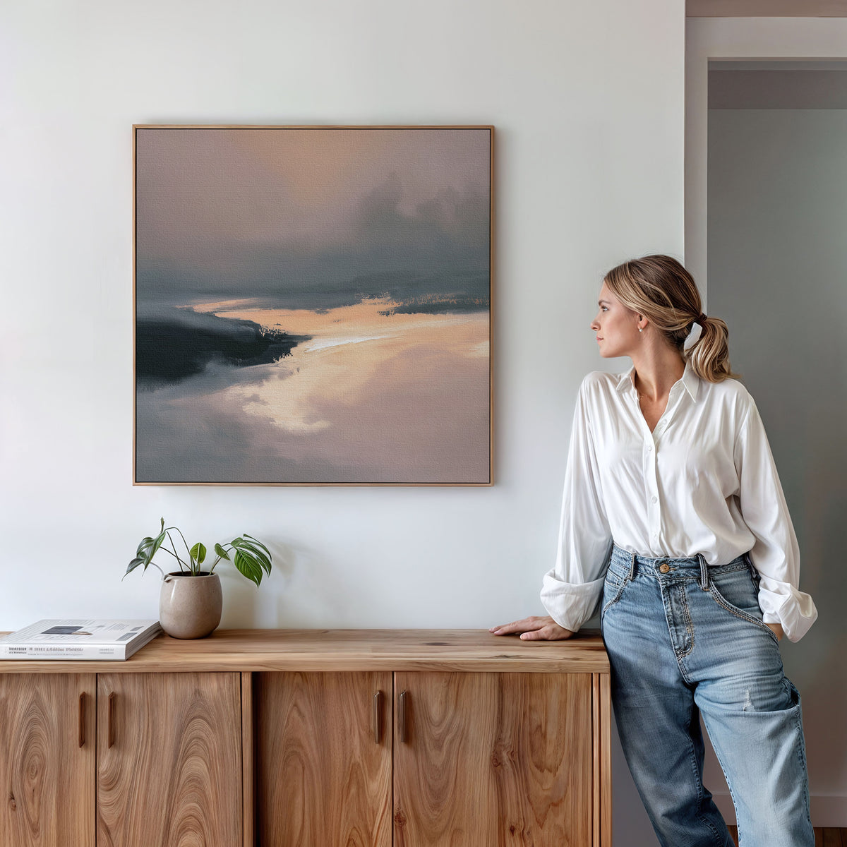 A woman in a white blouse and jeans stands by a wooden sideboard, admiring the Nordic Fjord Canvas Art - Square with blue, gray, and golden tones. A potted plant and books nearby complete this serene display.