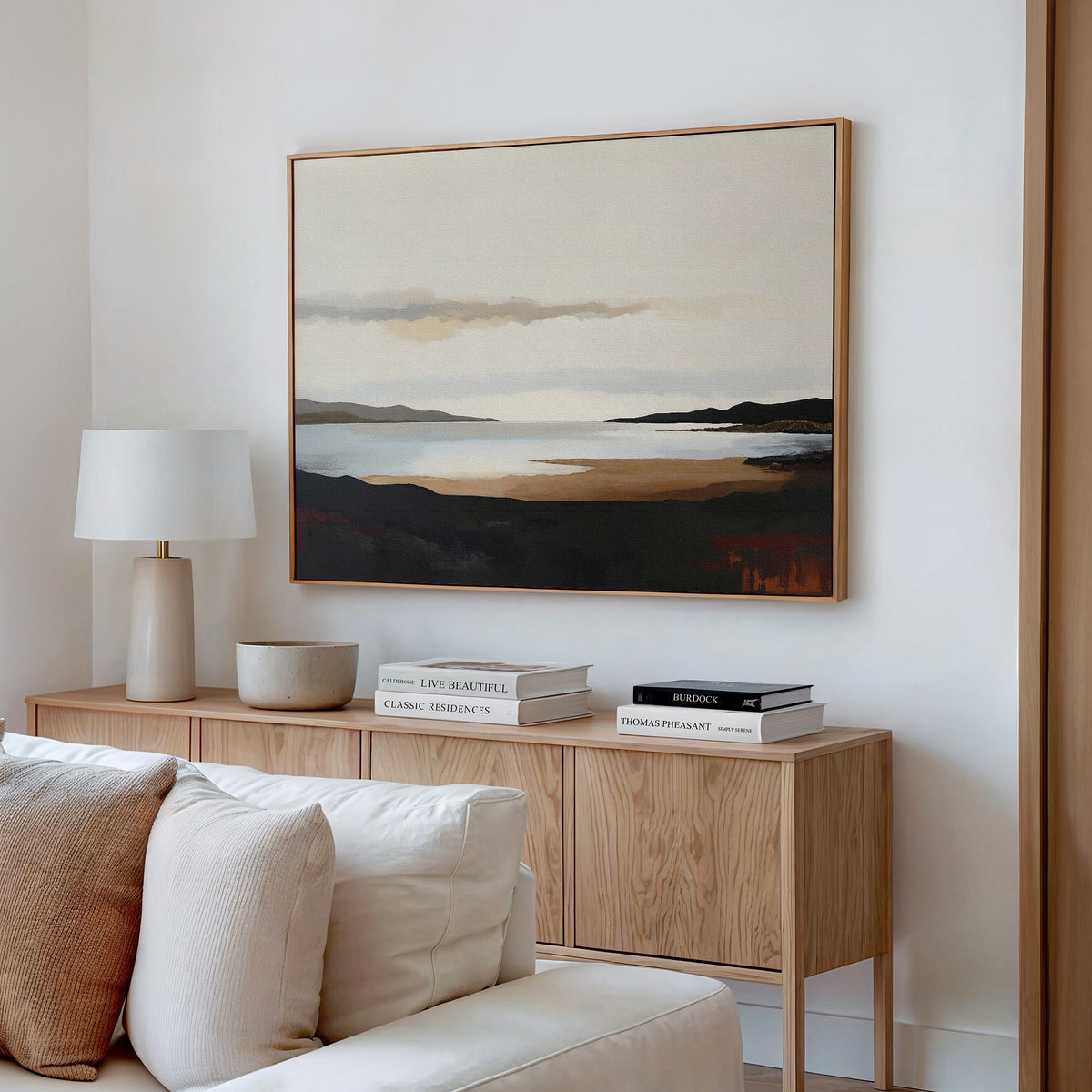 A modern living room has a light wood sideboard with books, a bowl, and a white lamp. Above it hangs the Nordic Waters Canvas Art - Horizontal, featuring mountains and water. In front sits a white sofa with cushions.