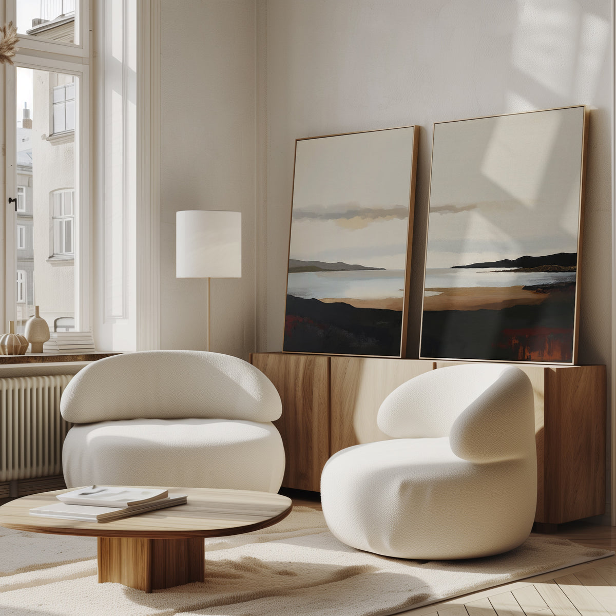 A modern living room with two curved white armchairs, a round wooden coffee table, and calming decor. The Nordic Waters - Set Of 2 landscape prints lean against the wall as large windows fill the space with natural light.