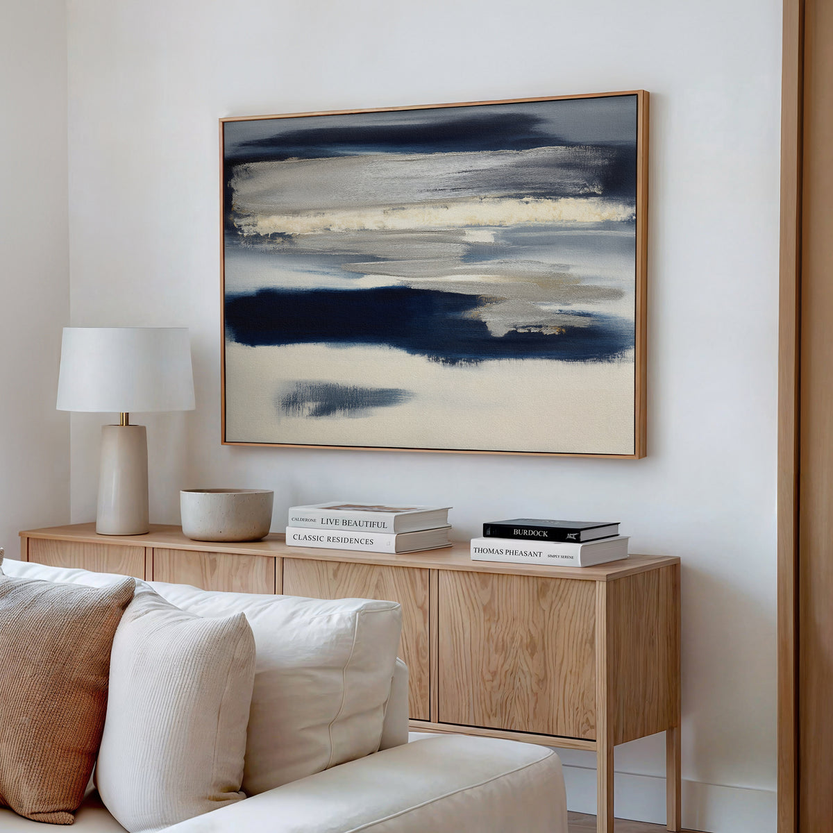 A modern living room features a light wood sideboard, white lamp, stacked books, neutral decor, and the Northern Tides Canvas Art - Horizontal—a striking abstract blue and beige painting—hanging above a cream sofa.