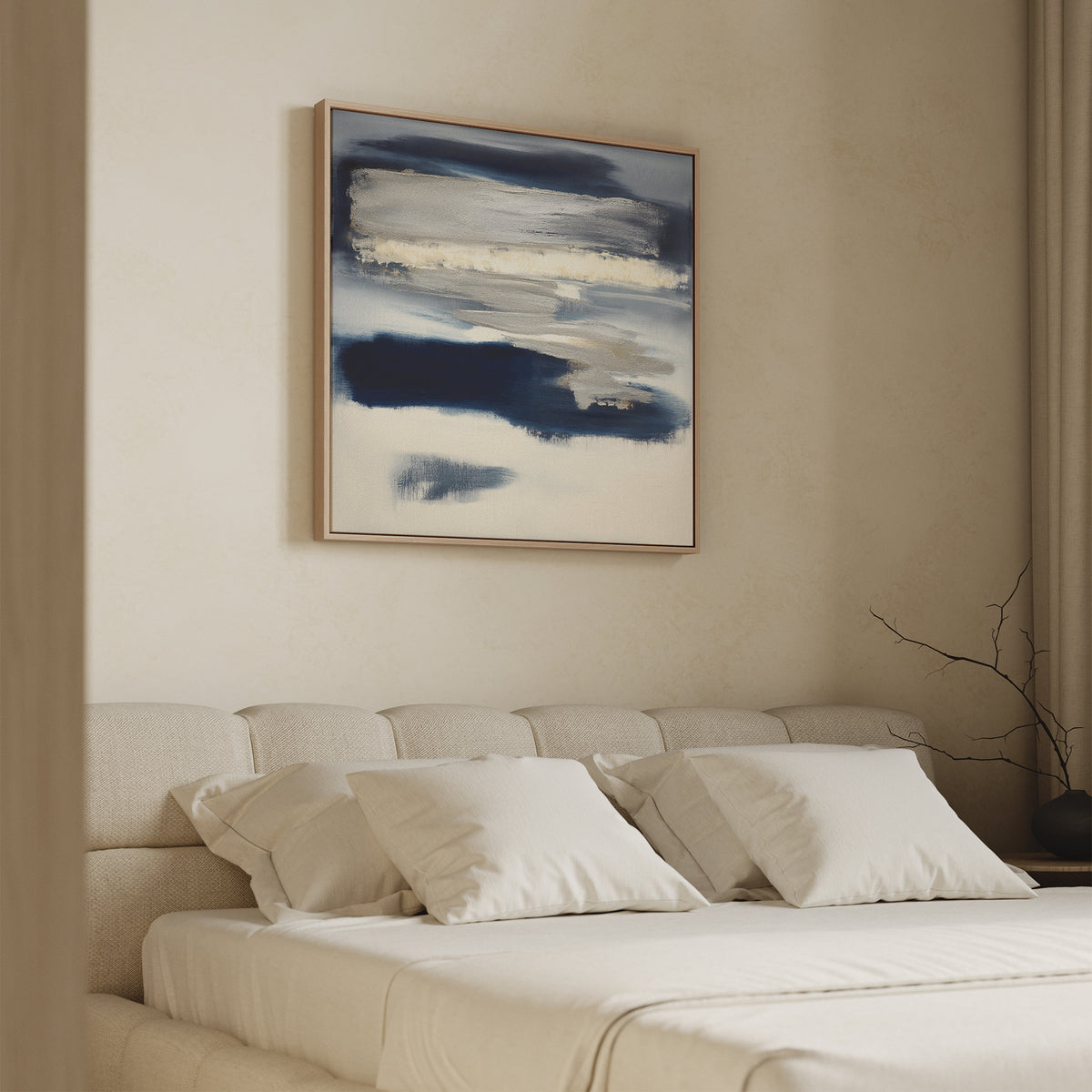 A modern bedroom features a beige upholstered bed with white bedding and pillows. Above the bed is the Northern Tides Canvas Art - Square, showcasing blue and white brushstrokes. A bare branch sits on the bedside table.