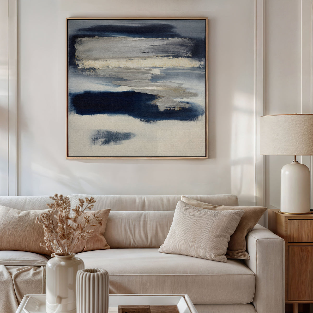 A modern living room with a beige sofa, neutral cushions, side table with ceramic lamp, and a vase of dried flowers. The Northern Tides Canvas Art - Square in indigo blue abstract style hangs above the sofa, adding a contemporary touch.