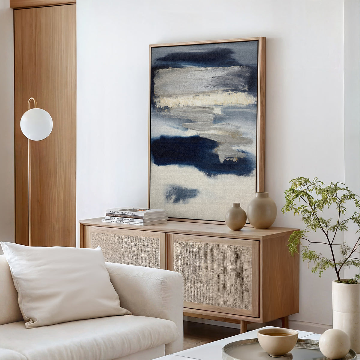 A modern living room with a light wood sideboard, Northern Tides Canvas Art - Vertical in an abstract print, neutral vases, a small green plant, a white sofa with cushions, and a round pendant light against minimalist white walls.