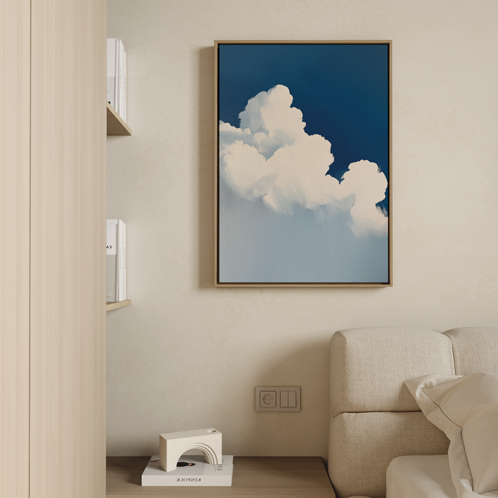 A modern living room with a beige sofa and light wood shelves features The Cumulus Abstract Canvas Art—minimalist wall art of white clouds on a blue sky—framed and displayed on a cream wall above an electrical outlet.