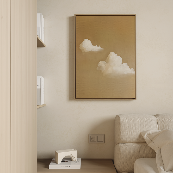 A minimalist living space features a beige sofa, shelves with white books, and the framed Whispering Clouds wall art depicting fluffy white clouds on a warm brown background hanging on a light-colored wall.