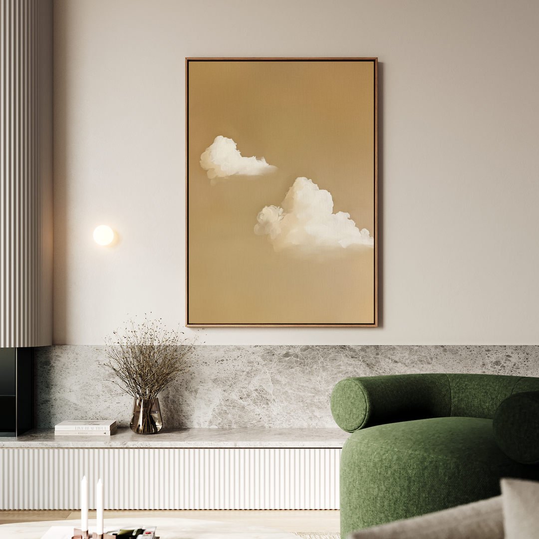 A modern living room showcases Whispering Clouds—a large, minimalist framed painting of white clouds on beige—mounted above a marble ledge with a glass vase of dried flowers, while a green armchair sits in the foreground.