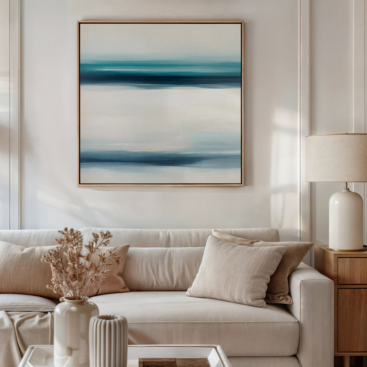 A modern living room features a beige sofa, light cushions, and a wooden side table with a lamp. A vase of dried flowers sits on the coffee table, while the Ocean Motion Canvas Art - Square above the sofa brings serene sophistication to the space.