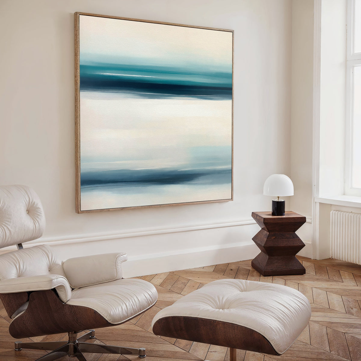 A modern living room with a white leather lounge chair, ottoman, geometric wooden side table with a white lamp, and the Ocean Motion Canvas Art - Square in blue and white tones creates an air of serene sophistication.
