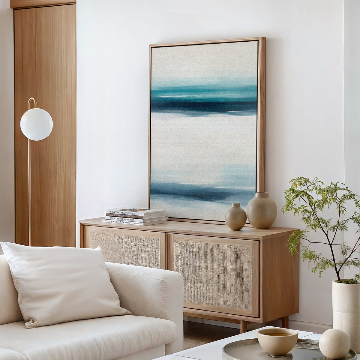 A modern, minimalist living room exudes serene sophistication with a light wood sideboard, ceramic vases, stacked books, a cream sofa, green plant, round pendant light, and the Ocean Motion Canvas Art - Vertical in blue and white.