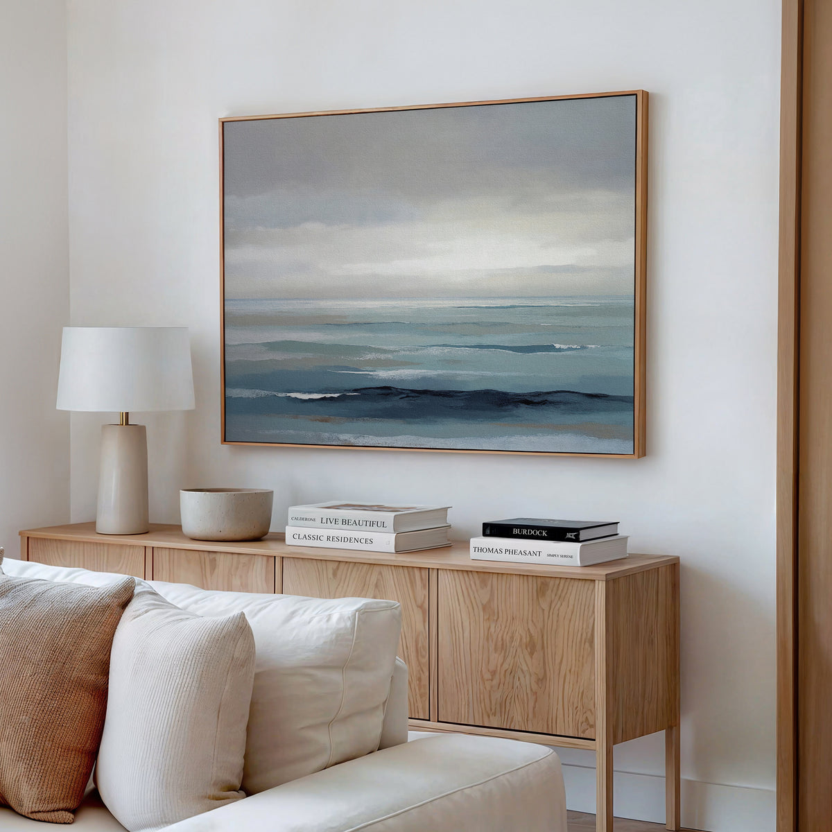 A modern living room features a wooden sideboard with stacked books, a white lamp, and a bowl. Above hangs the Ocean Swells Canvas Art - Horizontal, showcasing calm ocean hues of blue and gray. A white sofa sits in the foreground.
