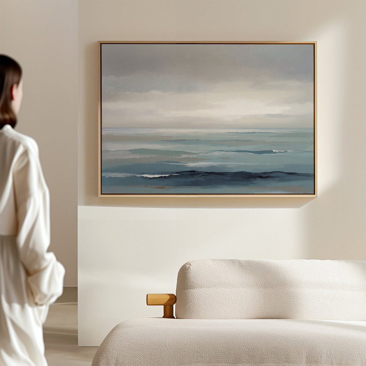A person in a white outfit stands in a minimal, light-filled room, gazing at the Ocean Swells Canvas Art - Horizontal—a coastal print in blue and gray tones—hanging above a textured cream-colored sofa.