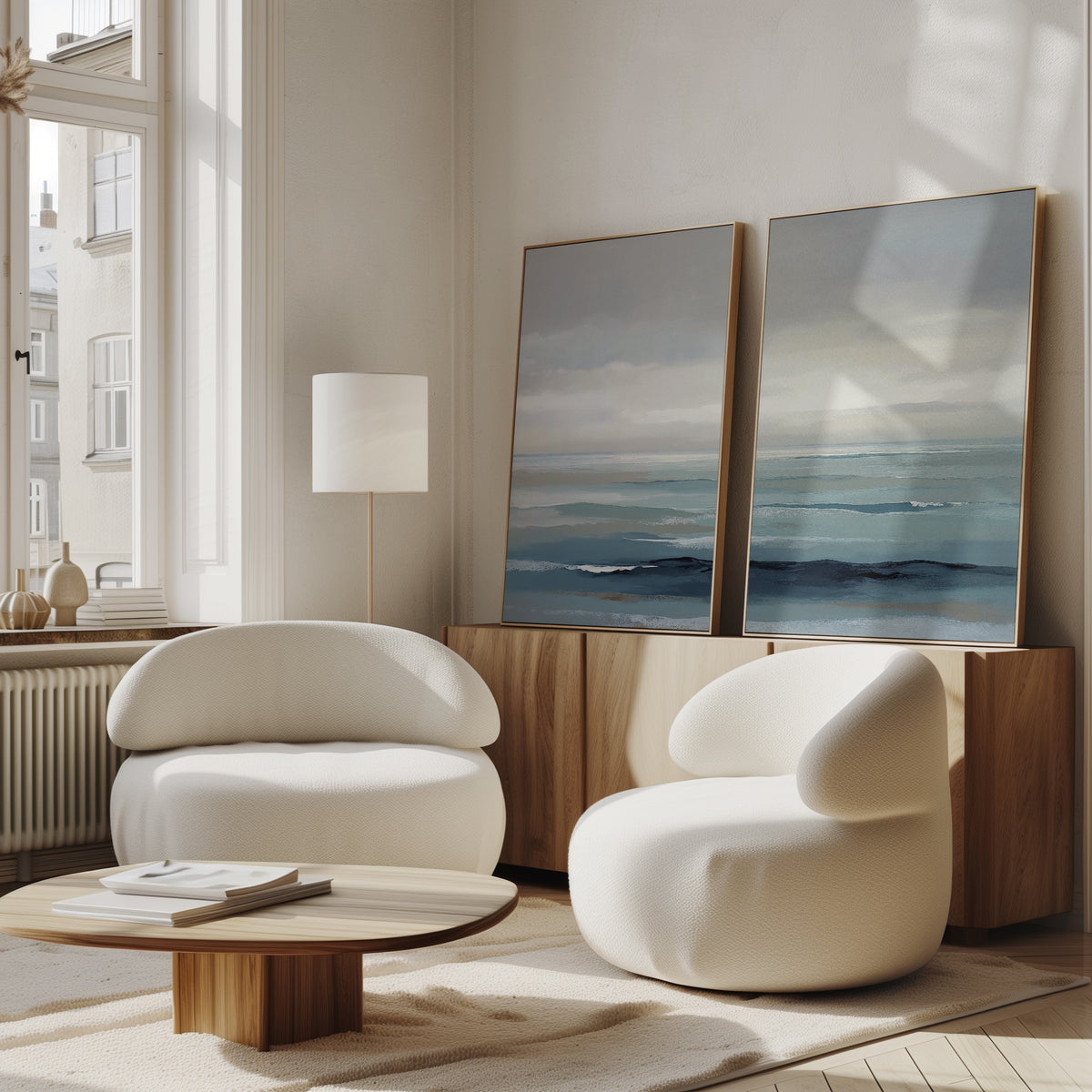 A modern living room with two white, rounded armchairs, a low wooden coffee table, a wooden sideboard topped with the Ocean Swells - Set Of 2 wall art; natural light brightens this Nordic Park-inspired space.