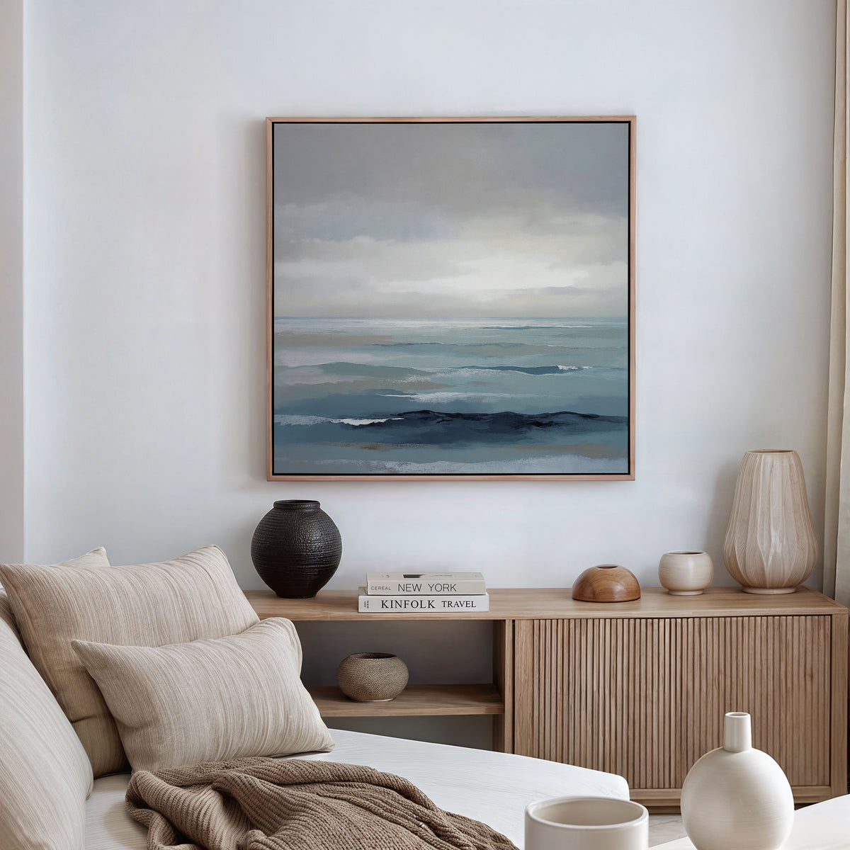 A minimalist living room features a beige sofa, neutral cushions, and a wooden sideboard. Above vases and books hangs the Ocean Swells Canvas Art - Square, infusing Nordic Park calm as natural light streams through the curtain.