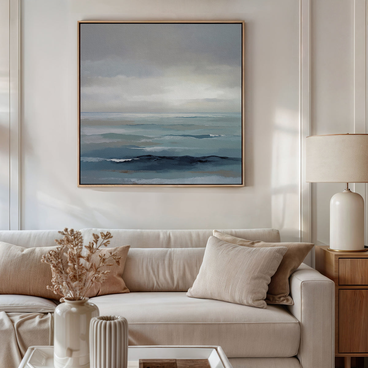 A modern living room with a white sofa, beige cushions, a wooden side table topped with a white lamp, and Ocean Swells Canvas Art - Square on the wall. A vase of dried flowers decorates the coffee table in front of the sofa.