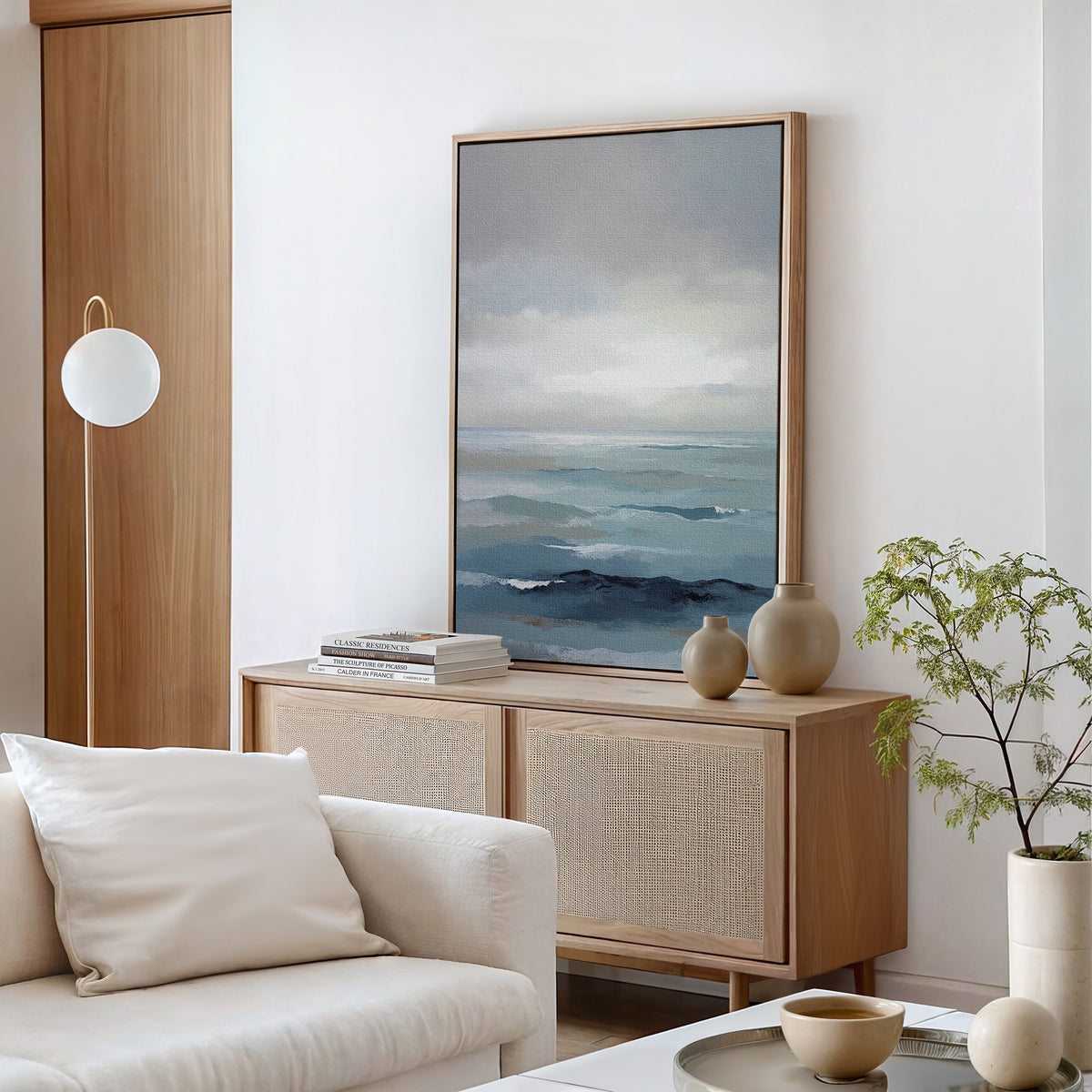 A modern living room with a light wood console, neutral decor, stacked books, ceramic vases, and a potted plant. The "Ocean Swells Canvas Art - Vertical" hangs on a white wall behind a white sofa with cushions.